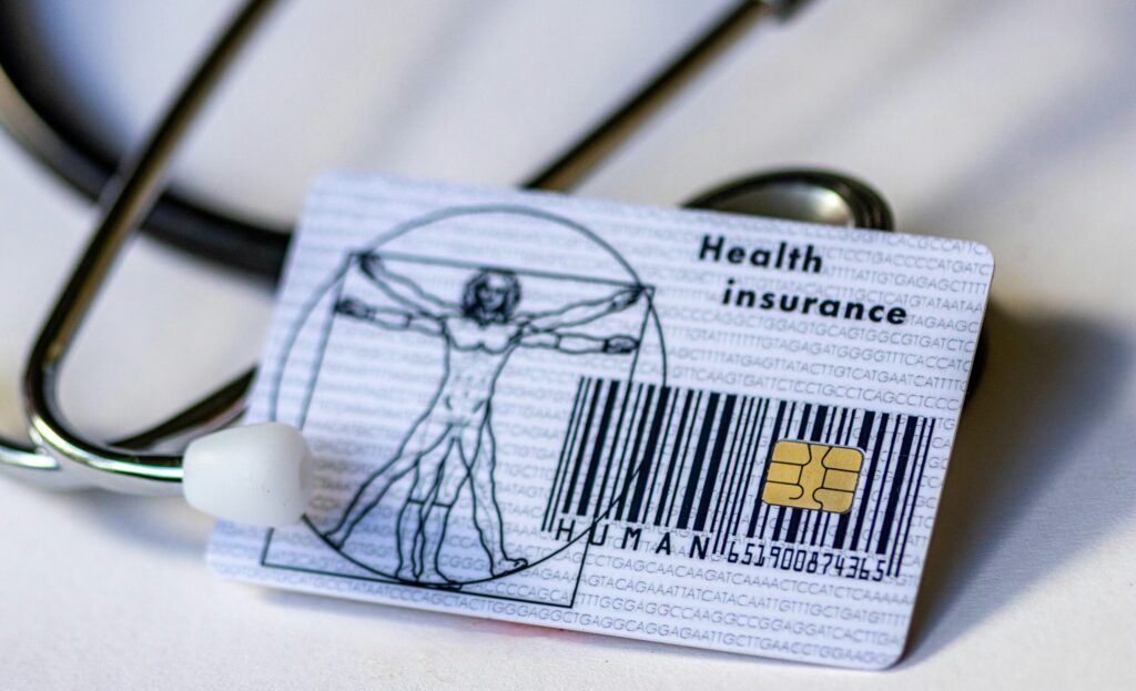 A close-up of a health insurance card placed on a white surface with a stethoscope partially visible in the background. The card features an illustration of Leonardo da Vinci’s Vitruvian Man on the left side, a gold chip for electronic use on the right, and a barcode below. The background of the card is filled with repeated sequences of DNA base letters (A, T, C, G). The words "Health insurance" are printed in the top right corner of the card.