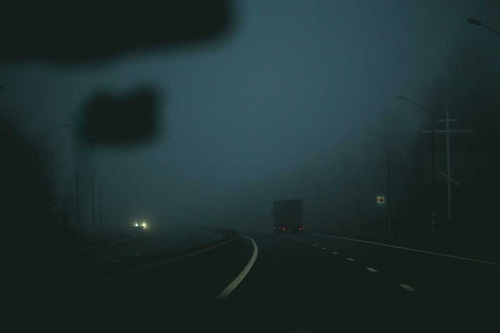 A dark, foggy highway at night, viewed from inside a vehicle. A large truck with red taillights is driving ahead in the right lane, barely visible through the thick fog. Another vehicle with bright headlights approaches from the opposite direction. Streetlights line both sides of the road, and a large white cross stands on the right side, possibly a roadside memorial or religious marker. The atmosphere is eerie and quiet, with limited visibility due to the dense fog.