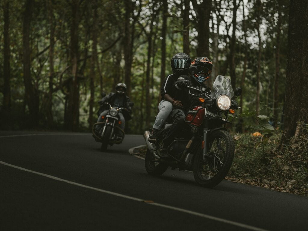 Two motorcyclists ride through a winding forest road. The rider in the foreground is carrying a passenger, both wearing helmets and protective gear. The motorcycle has a windshield and its headlight is on. Another motorcyclist follows behind, leaning into the curve. The scene is dimly lit by natural light filtering through tall, dense trees, creating a moody and adventurous atmosphere.