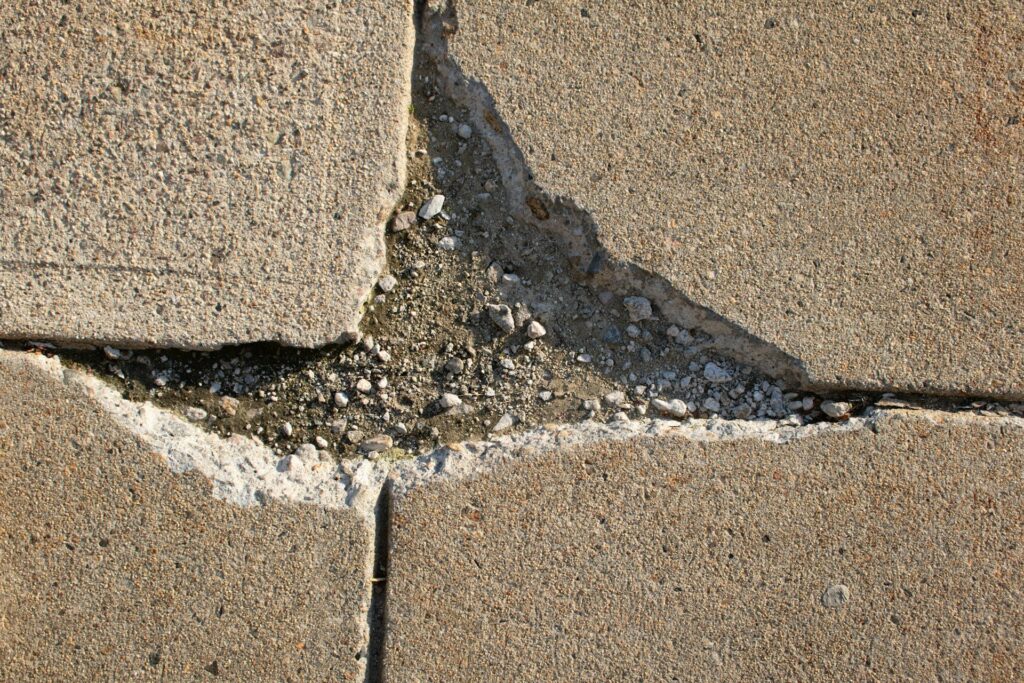 A close-up view of cracked and broken concrete pavement with loose gravel and debris filling the gaps between the slabs. The damage forms a jagged, uneven surface that poses a clear risk to pedestrian safety, especially for individuals with mobility issues or those using assistive devices like wheelchairs or canes.