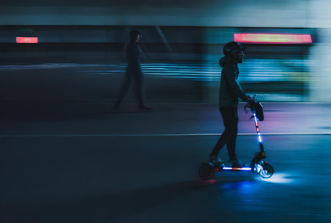 A person wearing a helmet rides an illuminated electric scooter through a dimly lit urban street at night. The scooter’s lights glow blue and red, leaving motion streaks. Another blurred pedestrian walks in the background.
