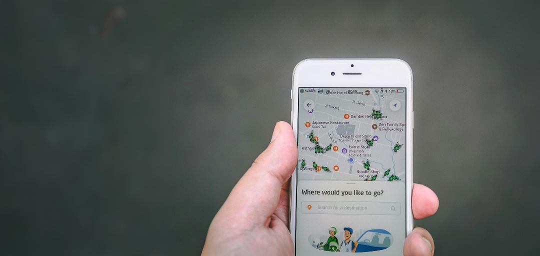 A close-up of a hand holding a white smartphone displaying a navigation app. The screen shows a city map with icons for shops, restaurants, and other destinations, along with a search bar that reads “Where would you like to go?” At the bottom of the screen are illustrated characters suggesting travel or rideshare options. The background is blurred and gray, keeping focus on the phone.