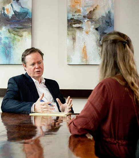 Tom McPherson, wearing a dark pinstripe suit and white shirt, sits at a polished wooden table speaking with a client. He gestures with his hands while holding a pen, emphasizing his points. The client, a woman with long hair wearing a dark blouse, is seated across from him with her back to the camera. Two abstract paintings hang on the wall behind them.