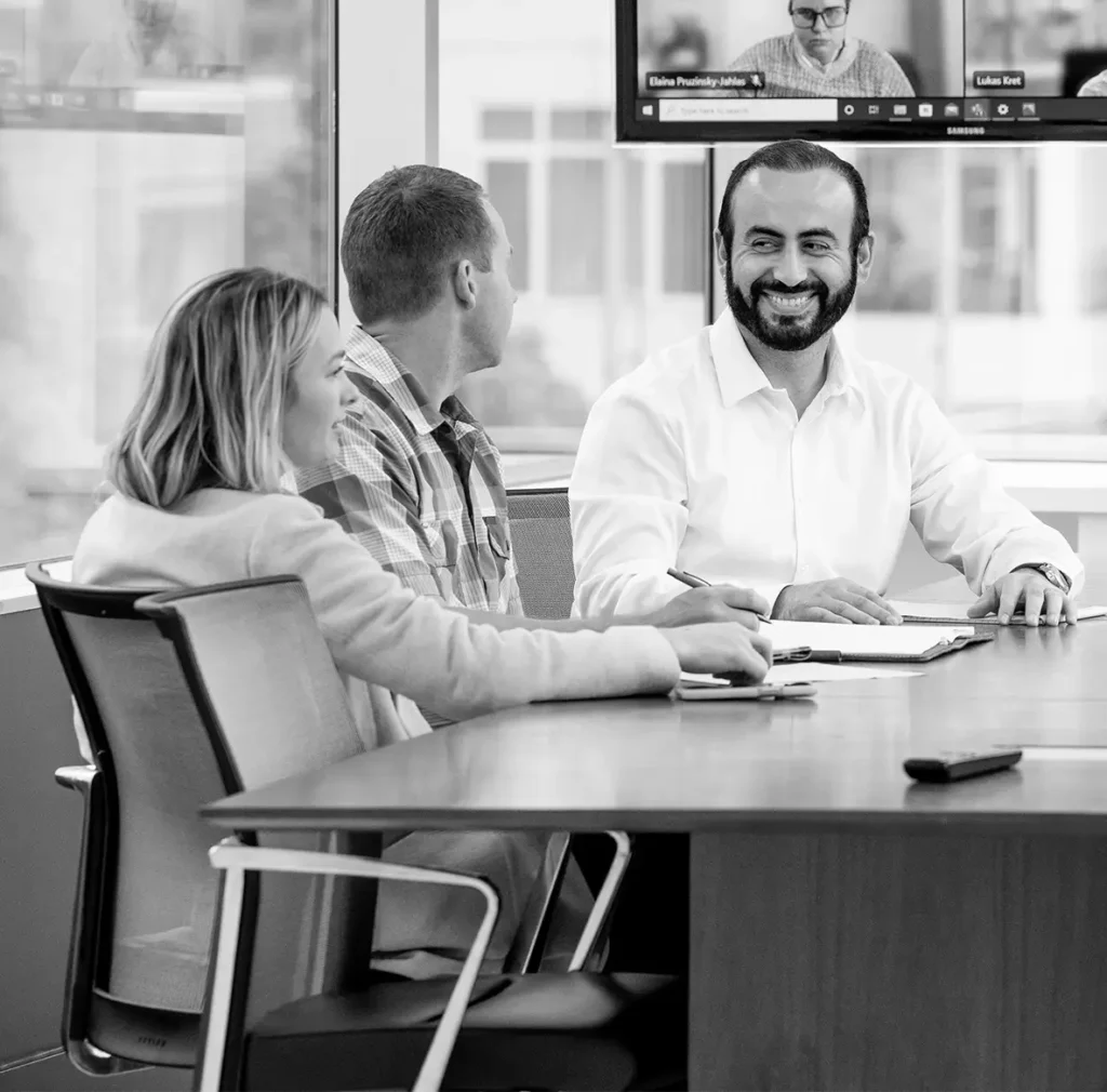 Black-and-white image of a group seated at a conference table during a hybrid meeting.