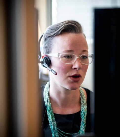 A person wearing glasses and a headset microphone is working at a desk, speaking while looking at a computer screen. They have short, styled hair brushed to the side and are wearing a black top with layered turquoise bead necklaces. The background shows an office environment with natural light.