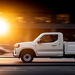 A white pickup truck drives quickly down a city street, captured in motion with a blurred background of cars and buildings. The sun is setting, casting a bright golden glow on the left side of the image, creating a dramatic light effect. The driver is visible through the window, and the truck appears to be moving steadily forward, emphasizing speed and urban energy.