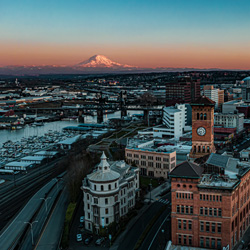 An aerial view of downtown Tacoma, Washington, during sunset. The city is filled with historic brick buildings, including a tall clock tower in the center, surrounded by modern structures. To the left, a marina with rows of boats sits along the water, and train tracks run parallel to the harbor. In the distance, Mount Rainier rises majestically with its snow-covered peak glowing pink from the setting sun, framed by a clear blue sky fading into warm orange tones near the horizon.