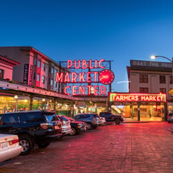 A brightly lit neon sign reading “Public Market Center” glows above the entrance to Seattle’s famous Pike Place Market at dusk. The words “Farmers Market” are also lit up in red on a nearby building. The market is surrounded by older-style buildings, with several parked cars lining the cobblestone street in front. The sky is a clear deep blue as night approaches, and warm yellow light spills from shop windows, giving the scene a welcoming and iconic atmosphere.