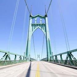 A symmetrical view of the St. Johns Bridge in Portland, Oregon, showing its iconic green Gothic-style towers and sweeping suspension cables. The image is taken from the center of the empty roadway, looking straight through the pointed arches of the tall towers, which rise high against a vibrant, cloudless blue sky. The double yellow line runs down the middle of the road, drawing the eye into the distance and emphasizing the bridge’s elegant structure and scale.