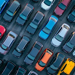 An overhead photo of a crowded parking lot filled with neatly lined rows of cars. The vehicles are parked closely together in alternating colors, including bright shades of red, yellow, orange, and blue mixed with black, white, and gray cars. The organized rows and variety of colors create a striking, patterned visual effect.