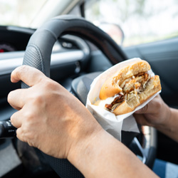 Close-up of a person’s hands inside a car. One hand is gripping the steering wheel, while the other holds a partially eaten sandwich wrapped in white paper. The sandwich appears to contain meat and sauce. The background shows a blurry view through the windshield, indicating that the car may be in motion. The image conveys the common but risky behavior of eating while driving.