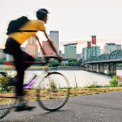 A blurred motion image of a person riding a bicycle on a paved path along the riverbank in Portland, Oregon. The cyclist, wearing a yellow shirt, black pants, a helmet, and a backpack, is in the foreground. Behind them is the iconic green truss Hawthorne Bridge spanning the Willamette River. The Portland city skyline, featuring various tall buildings, is visible across the water under a soft, pastel sky as the sun sets.