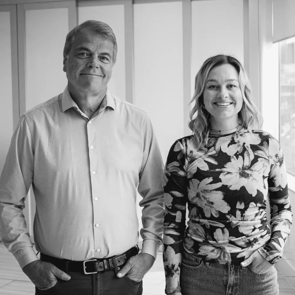 Black-and-white photo of Ken and Doni standing side by side indoors. Ken, on the left, is wearing a light button-up shirt with dark pants and has his hands in his pockets. Doni, on the right, is smiling and wearing a floral patterned long-sleeve top with jeans, also with one hand in her pocket. They are standing in front of a bright window and paneled wall.