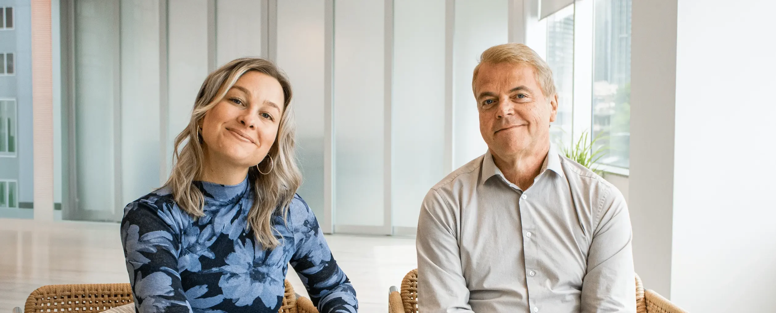 Doni sits cross-legged on a wicker chair, wearing a blue floral-patterned top, smiling toward the camera. Ken sits beside her in a light gray button-up shirt, also smiling. They are in a modern office with light walls, windows, and plants in the background.