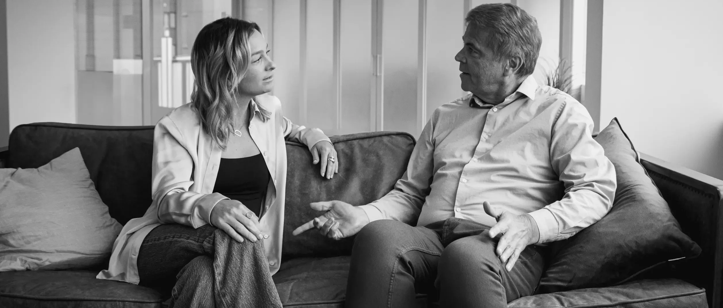 Black-and-white photo of Ken and Doni sitting on a couch in a modern office space. Ken, on the right, is wearing a light-colored button-up shirt and dark pants, gesturing with his hands while speaking. Doni, on the left, is wearing a light blouse over a dark top with jeans, sitting with her legs crossed and listening attentively. Cushions and a plant are visible in the foreground.