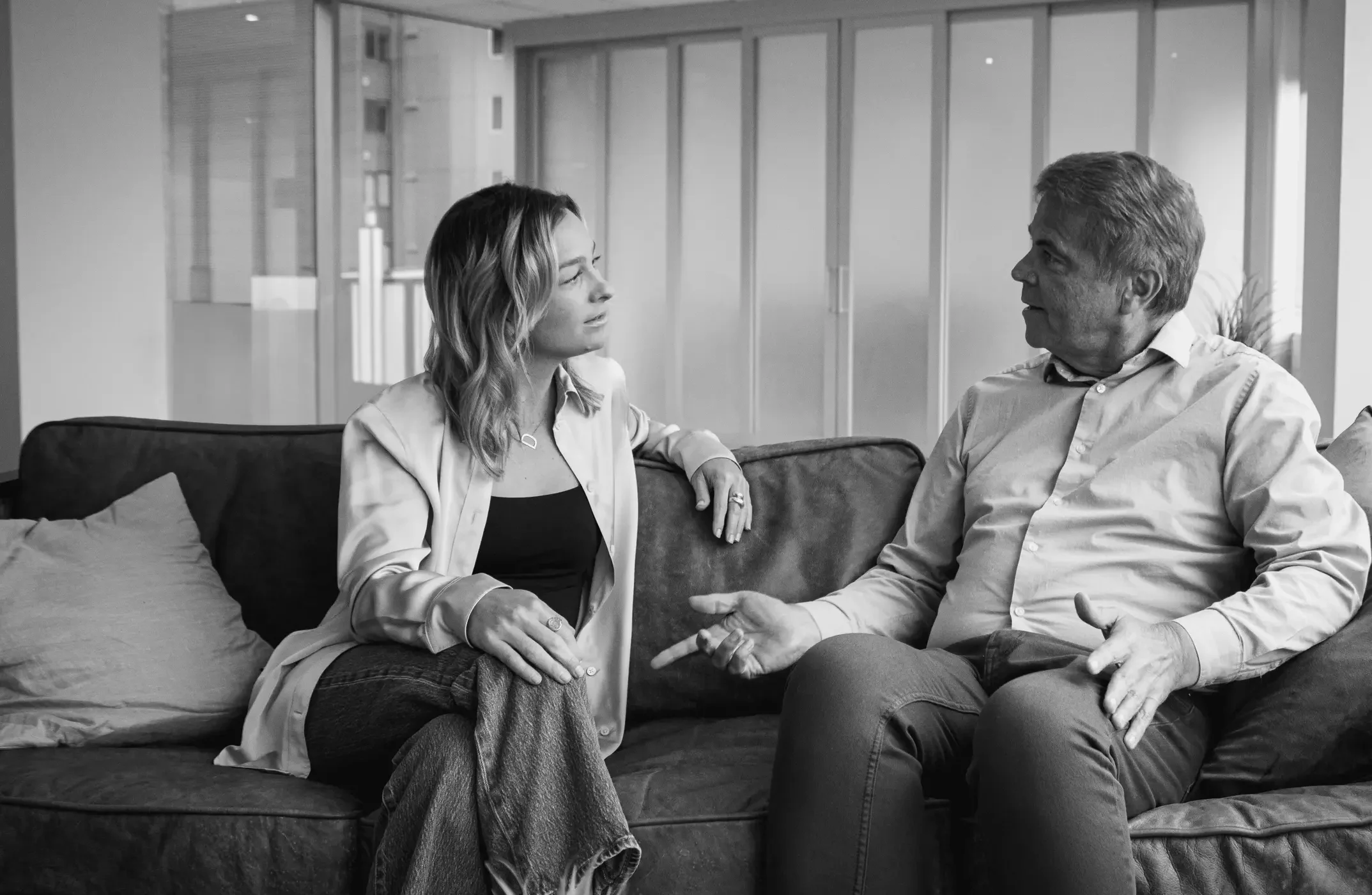 Black-and-white photo of Ken and Doni sitting on a couch in a modern office space. Ken, on the right, is wearing a light-colored button-up shirt and dark pants, gesturing with his hands while speaking. Doni, on the left, is wearing a light blouse over a dark top with jeans, sitting with her legs crossed and listening attentively. Cushions and a plant are visible in the foreground.