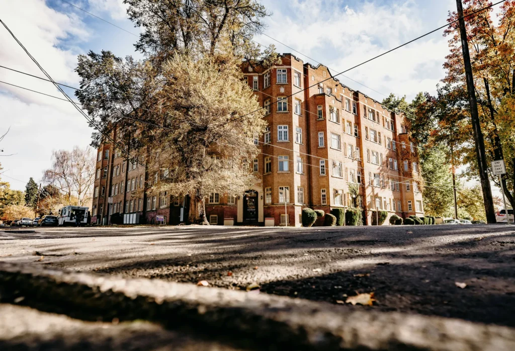 A wide-angle street-level view of a large historic brick apartment building with white-trimmed windows, surrounded by tall trees with autumn leaves. Shadows stretch across the pavement under a bright, partly cloudy sky.