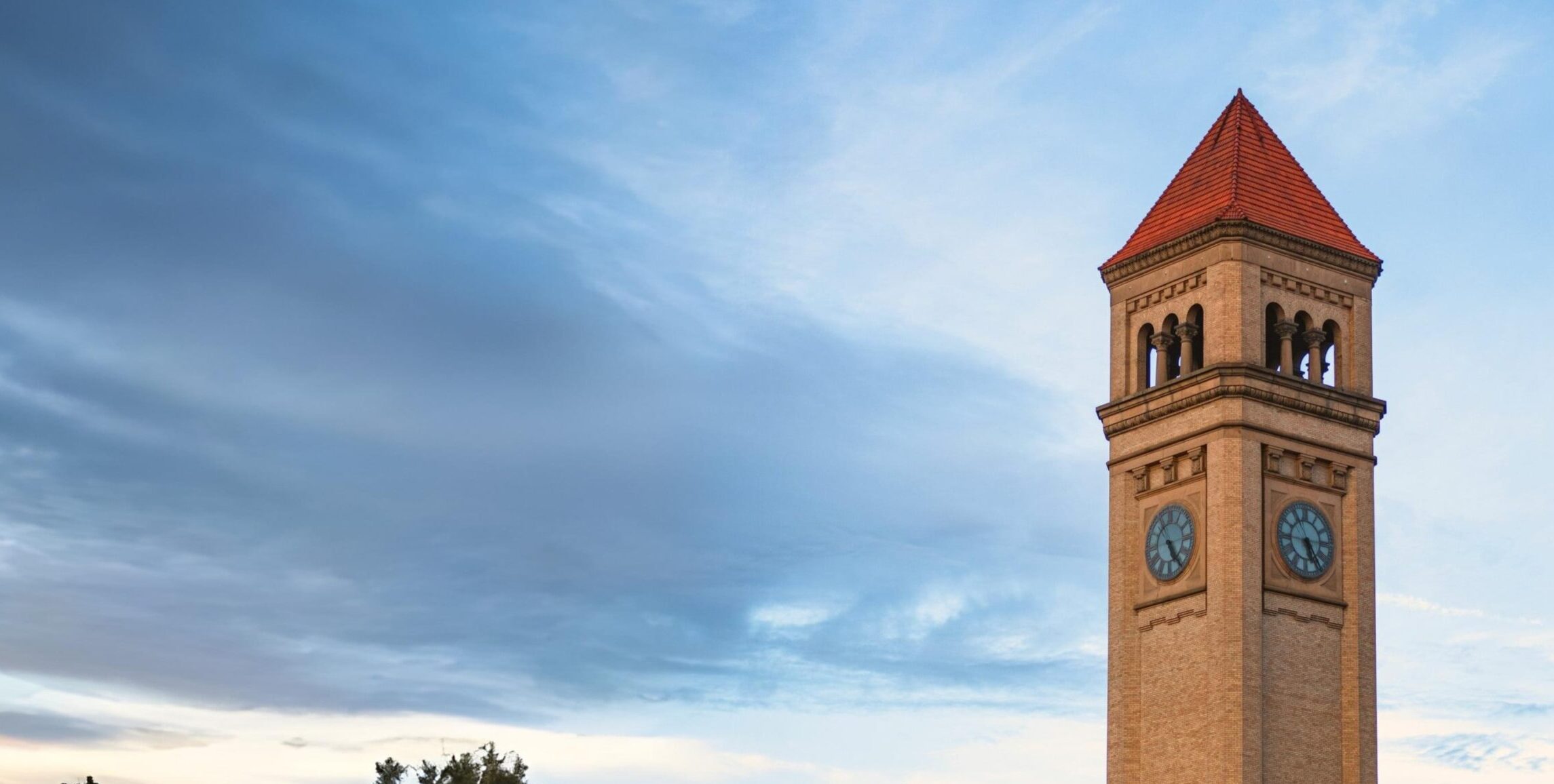 A tall brick clocktower with a red-tiled roof rises above the trees under a blue and cloudy sky. This landmark stands across the street from our Spokane office, where our pedestrian accident attorney team helps clients with crosswalk and roadway safety cases.