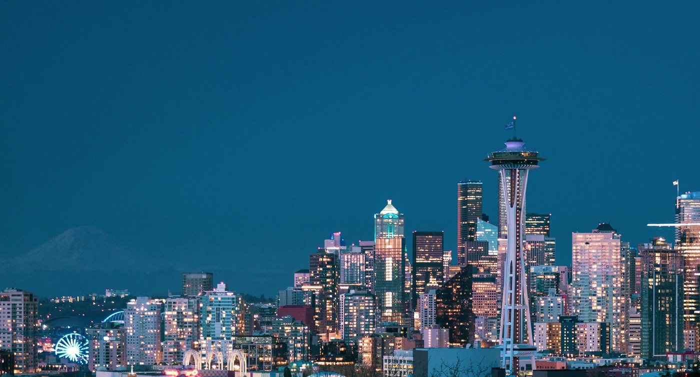 A twilight view of downtown Seattle with the iconic Space Needle on the right side of the image. The skyline is lit with glowing office and residential building lights against a deep blue evening sky. Mount Rainier is faintly visible in the background on the left, blending into the horizon. The cityscape captures both modern skyscrapers and the landmark tower, symbolizing Seattle’s vibrant urban atmosphere.