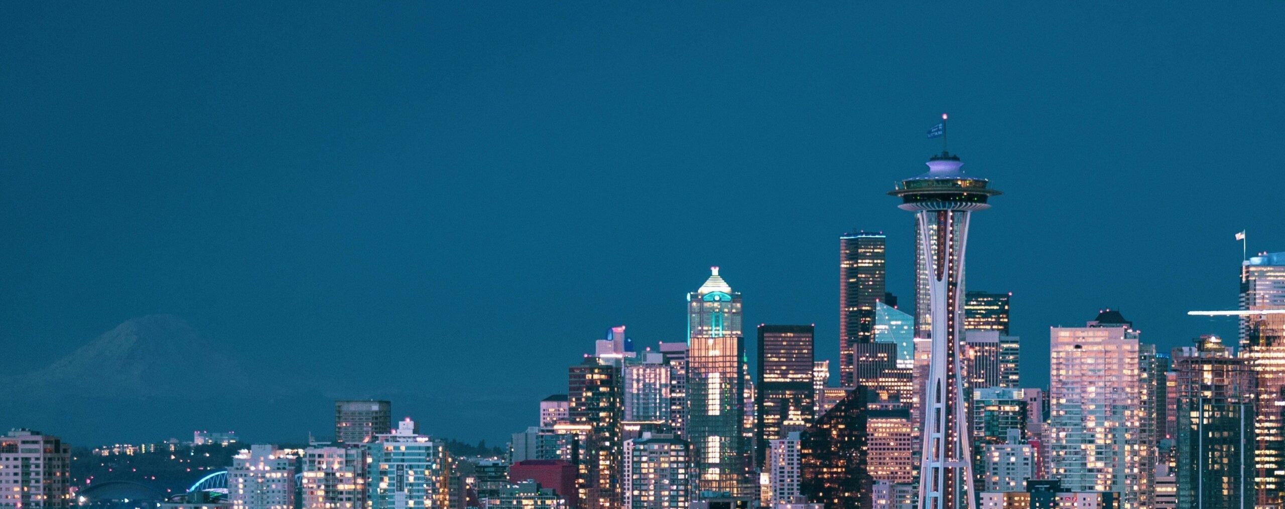 A twilight view of downtown Seattle with the iconic Space Needle on the right side of the image. The skyline is lit with glowing office and residential building lights against a deep blue evening sky. Mount Rainier is faintly visible in the background on the left, blending into the horizon. The cityscape captures both modern skyscrapers and the landmark tower, symbolizing Seattle’s vibrant urban atmosphere.