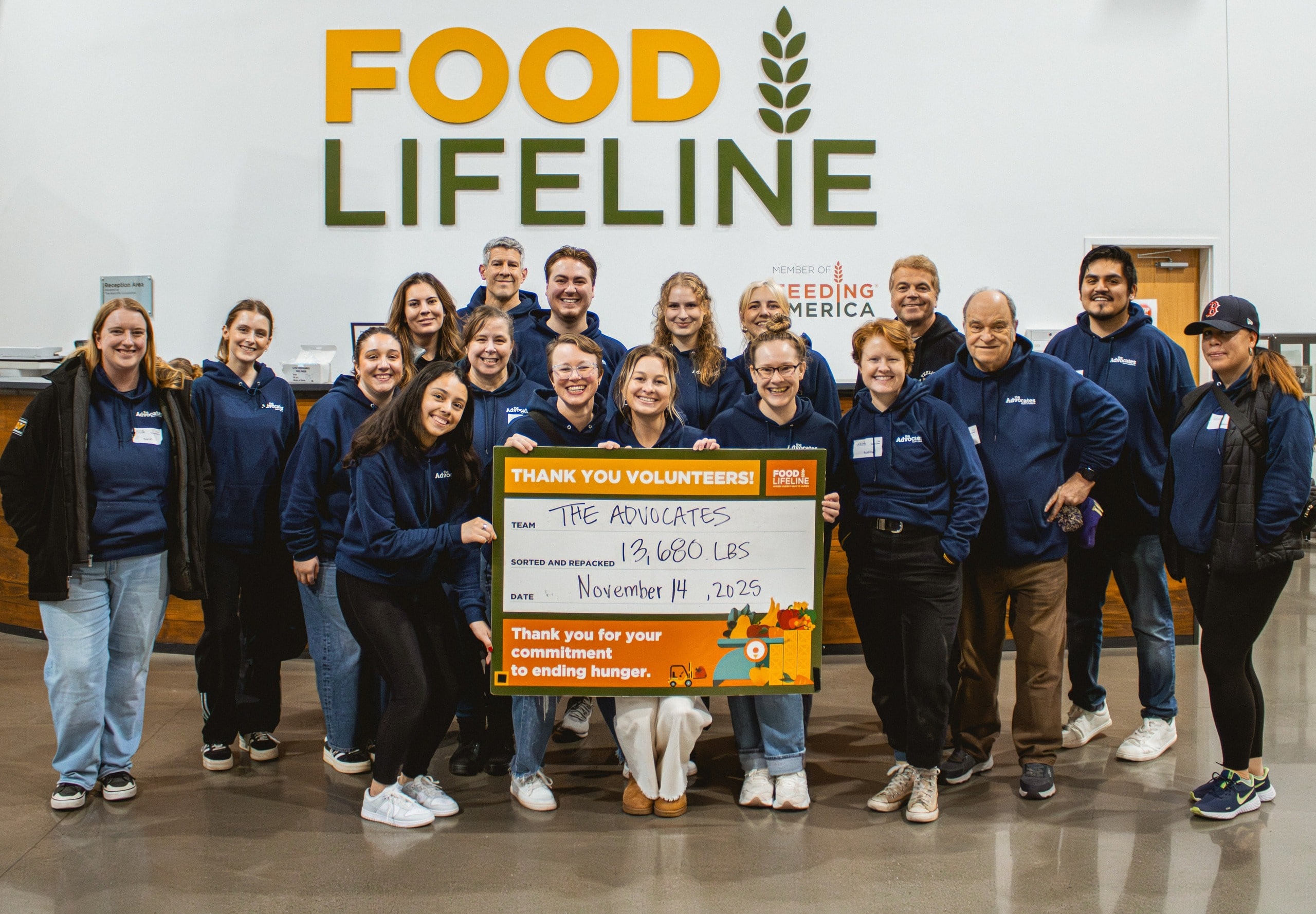 A group of The Advocates Injury Attorneys staff pose together inside the Food Lifeline facility, smiling and holding a large orange and white sign that reads, “Thank you volunteers! Team: The Advocates, 13,680 lbs sorted and repacked, Date: November 14, 2025.” Everyone is wearing matching navy blue hoodies with The Advocates logo, standing in front of a wall displaying the Food Lifeline logo and the Feeding America emblem.